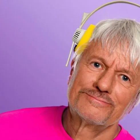 Ein älterer Mann mit grauen Haaren trägt gelbe Kopfhörer. Er hat ein freundliches Lächeln und trägt ein pinkes T-Shirt.