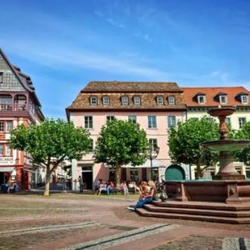 Ein schöner Platz mit bunten Gebäuden und Bäumen. In der Mitte befindet sich ein Brunnen, umgeben von Menschen, die die Sonne genießen.