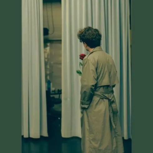 Ein Mann in einem langen Mantel steht mit einer roten Rose in der Hand vor einem Vorhang. Er blickt nachdenklich in den Raum.
