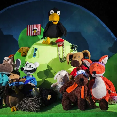 Eine bunte Bühne mit verschiedenen Plüschfiguren, darunter ein Waschbär, ein Hund und ein Fuchs. Im Hintergrund steht ein gelber Vogel und es gibt eine grüne Landschaft.