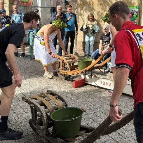Zwei Teilnehmer bei einem Wettrennen mit Schubkarren auf einer belebten Straße. Zuschauer beobachten gespannt das Geschehen.
