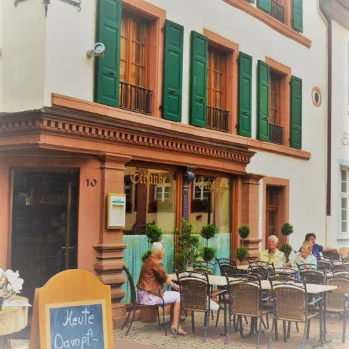 Ein gemütliches Café mit Tischen im Freien und Gästen, die sich entspannen. Das Gebäude hat grüne Fensterläden und einen einladenden Eingang.