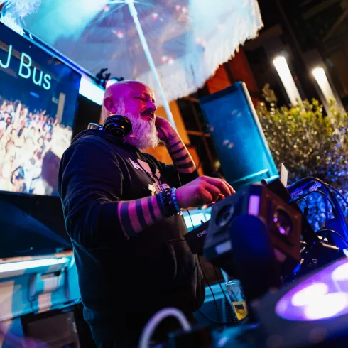 Ein DJ steht hinter einem Mischpult und spielt Musik. Im Hintergrund ist eine begeisterte Menge zu sehen, umgeben von bunten Lichtern.