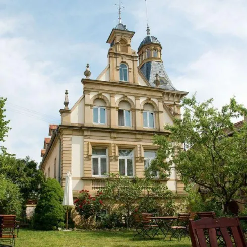 Ein elegantes historisches Gebäude mit einem grünen Garten. Die Architektur zeigt detailreiche Elemente und einen hohen Turm.