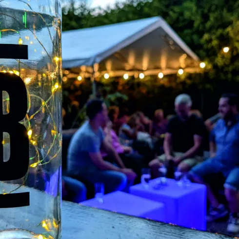 Ein festlicher Außenbereich mit Gästen, die sich unter Lichtern entspannen. Im Vordergrund steht eine Flasche mit dem Schriftzug „Weingut Buchert“.