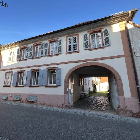 Ein historisches Gebäude mit weißen Wänden und blauen Fensterläden. Es hat einen durchgehenden Torbogen, der einen Weg zum Innenhof führt.