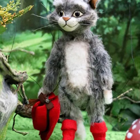 Eine marionettenartige Katze trägt rote Stiefel und hält einen roten Hut in der Hand. Sie steht in einer grünen Waldumgebung mit Pflanzen und Pilzen.