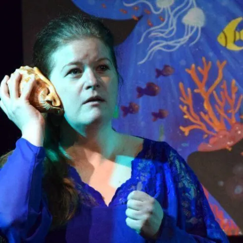 Eine Frau in einem blauen Outfit hält eine Muschel ans Ohr. Im Hintergrund ist ein buntes Wandgemälde mit Fischen und Korallen zu sehen.