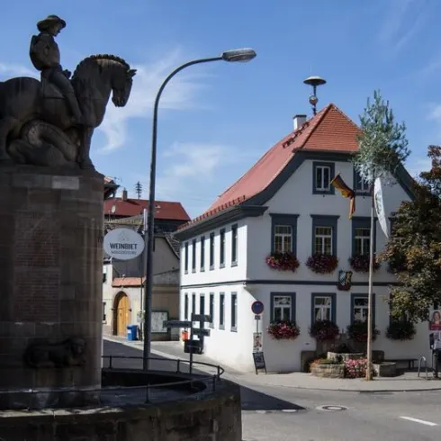 Eine charmante Stadtansicht mit einem Denkmal eines Reiters und einem historischen Gebäude im Hintergrund. Der Himmel ist klar und blau, und die Straßen sind mit Bäumen gesäumt.