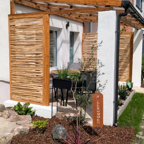 Ein gemütlicher Außenbereich mit Holzlatten und einem Tisch mit Stühlen. Eine Willkommens-Säule steht im Garten, umgeben von Pflanzen.