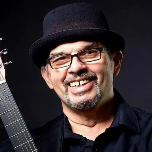 Ein lächelnder Mann mit einer Gitarre in der Hand trägt einen schwarzen Hut und eine Brille. Der Hintergrund ist dunkel und hebt seine positive Ausstrahlung hervor.