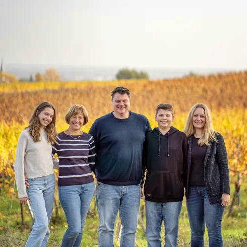 Eine Familie steht lachend in einem Weinberg mit herbstlichen Farben. Im Hintergrund sind die Reben und ein klarer Himmel zu sehen.