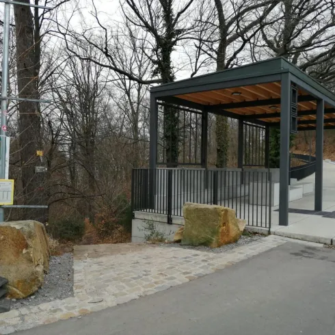 Eine moderne überdachte Haltestelle in einem Waldgebiet. Im Hintergrund sind kahle Bäume zu sehen, und der Boden ist gepflastert.