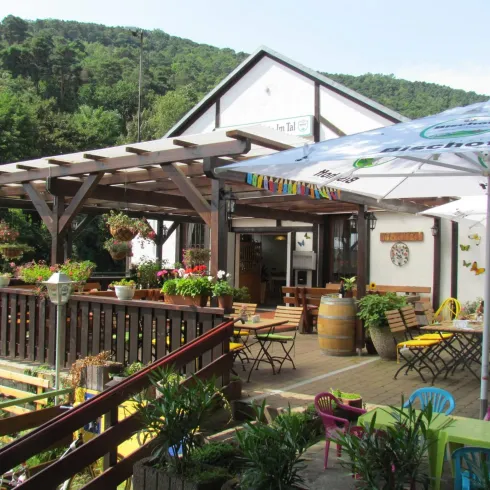 Ein einladendes Restaurant mit einer überdachten Terrasse und bunten Tischen. Der Außenbereich ist mit Pflanzen und Blumen geschmückt.