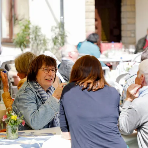 Eine Gruppe von Menschen sitzt draußen an Tischen und unterhält sich. Es scheint eine freundliche und gesellige Atmosphäre zu sein.