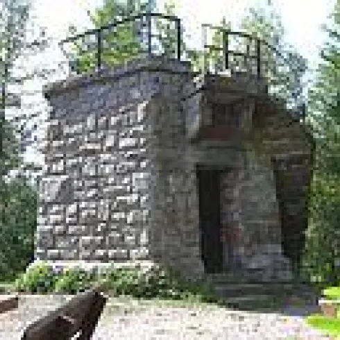 Ein historischer Steinturm umgeben von Bäumen. Er hat eine Aussichtsplattform und eine Eingangstür.