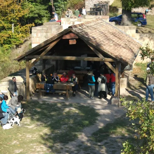 Ein Holzpavillon umgeben von Bäumen, in dem viele Menschen sitzen und sich entspannen. Es ist ein sonniger Tag und die Atmosphäre ist freundlich.