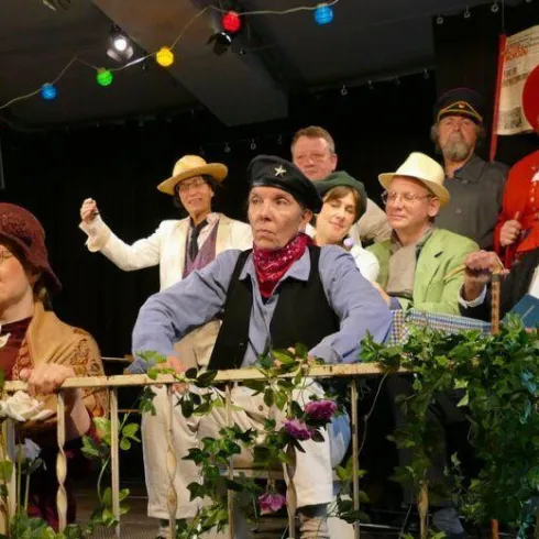 Eine Gruppe von Menschen steht und sitzt auf einer Bühne, gekleidet in bunte Kostüme. Sie scheinen an einer Theateraufführung teilzunehmen und lachen oder posieren für das Foto.
