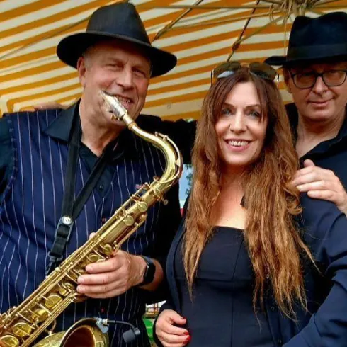 Drei Musiker stehen unter einem bunten Sonnenschirm. Ein Saxophonist, eine Sängerin und ein weiterer Mann posieren lächelnd für das Foto.