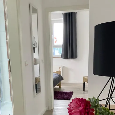 Ein moderner Flur mit einem Spiegel und einer großen Lampe. Im Hintergrund ist ein Schlafzimmer mit einem Bett und Vorhängen zu sehen.