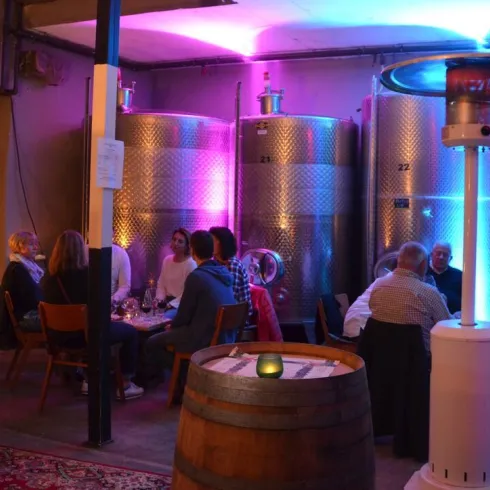 Eine gemütliche Weinbar mit Gästen, die an einem Tisch sitzen. Im Hintergrund stehen große silberne Weinfässer, beleuchtet von bunten Lichtern.