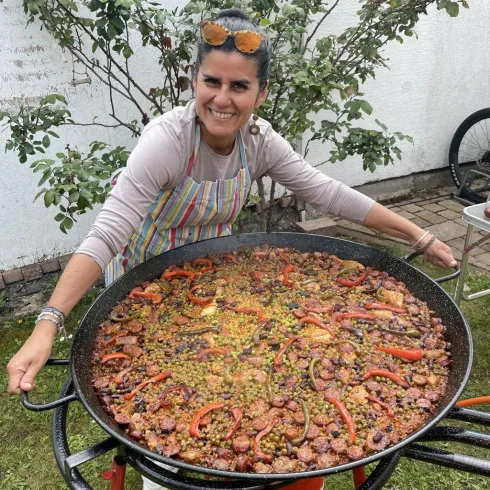 Eine Frau lächelt stolz und hält eine große Paella-Pfanne mit buntem Gemüse und Fleisch. Im Hintergrund sind Pflanzen und ein Fahrrad zu sehen.