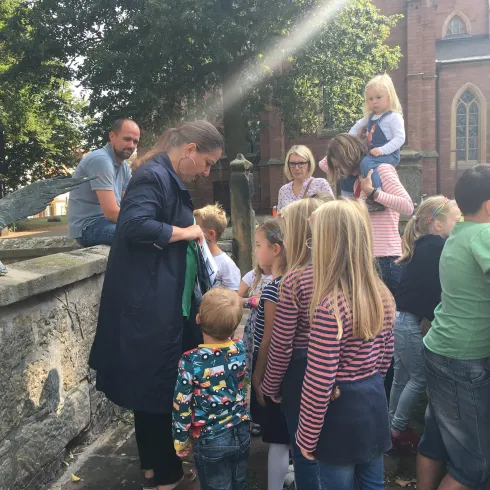 Eine Gruppe von Kindern beobachtet aufmerksam einen Erwachsenen, der etwas erklärt. Im Hintergrund ist eine alte Kirche und viel grüne Vegetation sichtbar.
