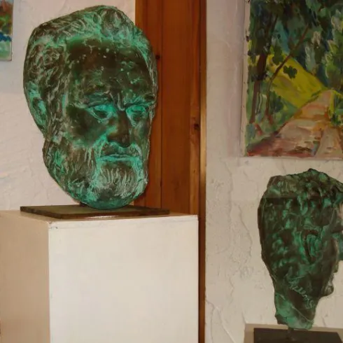 Zwei Skulpturen von Köpfen aus Bronze stehen in einer Galerie. Im Hintergrund sind bunte Gemälde zu sehen.