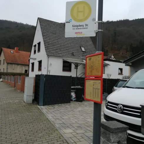 Eine Bushaltestelle mit einem klaren Schild an einer Straße. Im Hintergrund ist ein Wohnhaus und parkende Autos zu sehen.