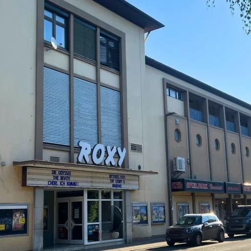 Ein modernes Kino mit dem Namen "Roxy" an einer straßenseitigen Lage. Die Fassade ist hell erleuchtet und es stehen Autos davor.