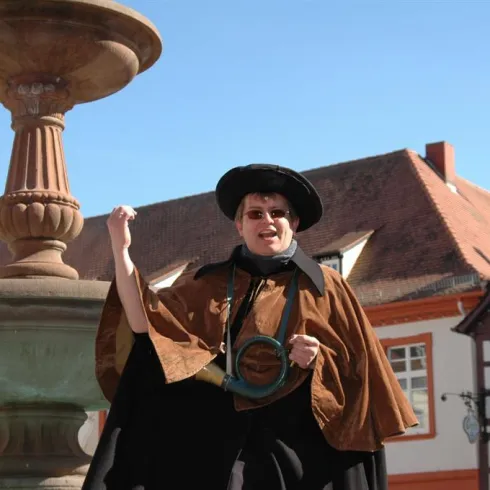 Eine Person in historischer Kleidung steht vor einem Brunnen und lächelt. Im Hintergrund sind traditionelle Gebäude mit roten Dächern zu sehen.