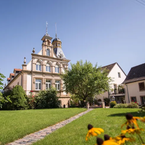 Ein historisches Gebäude mit Türmchen und einem schönen Garten. Im Vordergrund sind gelbe Blumen und ein gepflasterter Weg zu sehen.