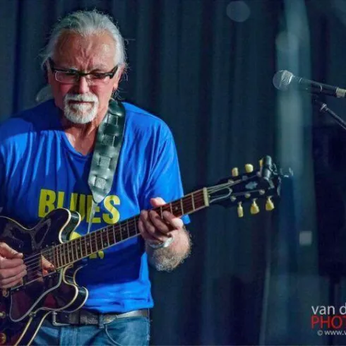 Ein Musiker spielt Gitarre auf der Bühne. Er trägt ein blaues T-Shirt mit der Aufschrift "BLUES" und hat weiße Haare.