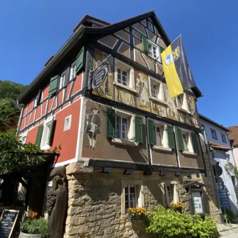 Ein historisches Fachwerkhaus mit bunten Wandmalereien und grünen Fensterläden. Darüber weht eine Flagge vor blauem Himmel.