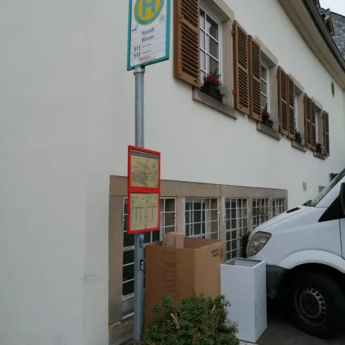 Eine Bushaltestelle mit einem Informationsschild steht neben einem weißen Transporter. Im Vordergrund sind Kartons und ein Topf mit einer Pflanze sichtbar.