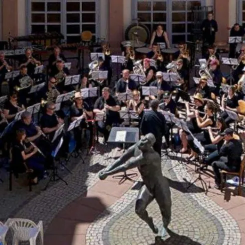 Ein Blasorchester spielt im Freien, umgeben von Zuschauern. Im Vordergrund steht eine Statue, die die Szenerie ergänzt.