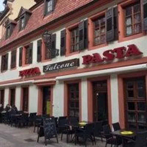 Ein italienisches Restaurant mit der Beschriftung „Pizzeria Falcone“ und „Pasta“. Es gibt Tische und Stühle im Freien.