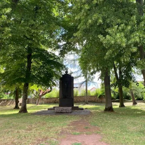 Ein Denkmal steht umgeben von hohen Bäumen in einem grünen Park. Der Platz wirkt ruhig und einladend.