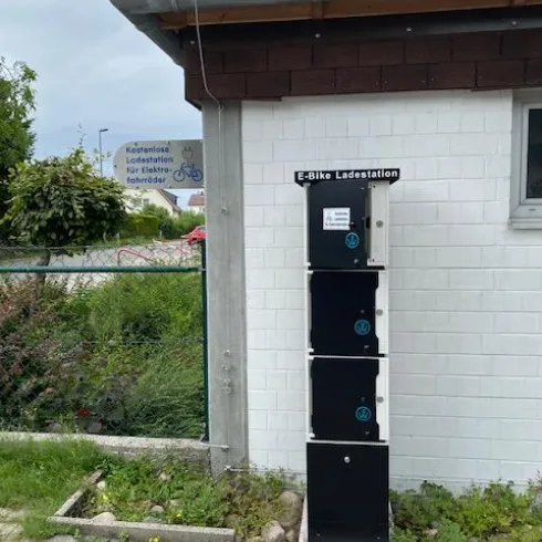 Eine E-Bike Ladestation mit mehreren Ladefächern steht an einer Wand. Im Hintergrund ist ein Schild mit Informationen zur E-Bike Ladestation zu sehen.