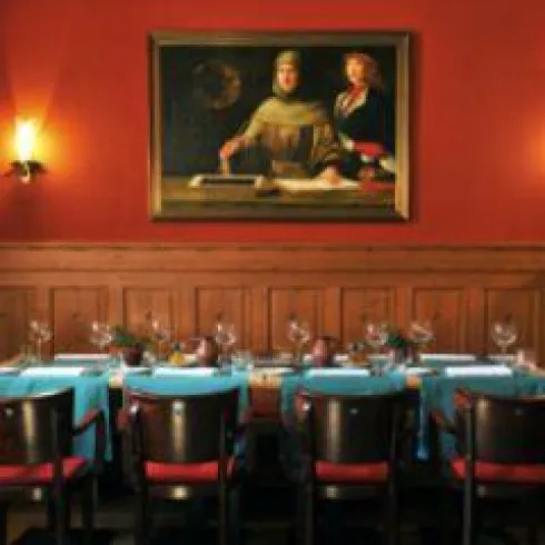 Ein elegantes Restaurant mit einem langen Tisch, der mit blauen Tischtüchern und Gläsern gedeckt ist. An der Wand hängt ein historisches Gemälde.