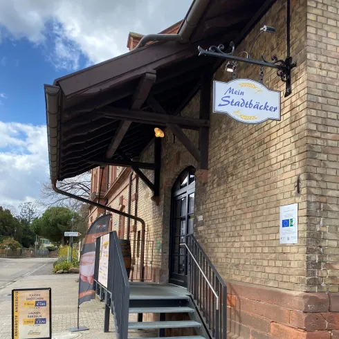Ein einladender Eingangsbereich mit einer Treppe und einem überdachten Vorbau. Über der Tür hängt ein Schild mit dem Namen "Mein Stadtbäcker".