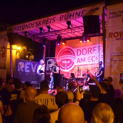 Eine lebendige Musikveranstaltung bei Nacht. Die Bühne ist mit bunten Lichtern beleuchtet und eine Menschenmenge genießt die Aufführung.