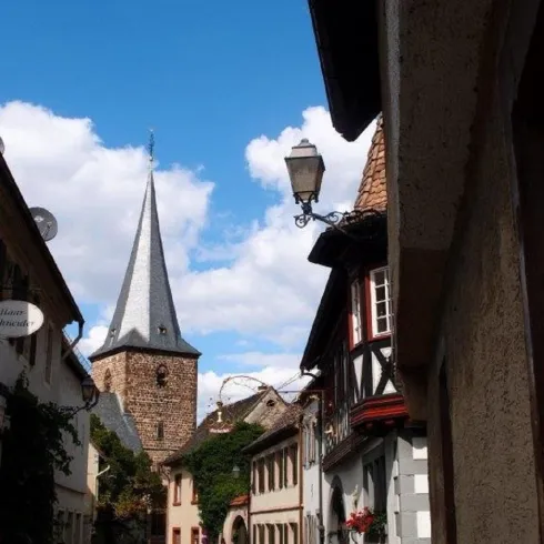 Eine malerische Straße in einem historischen Dorf mit alten Häusern. Im Hintergrund erhebt sich ein Kirchturm unter einem blauen Himmel.