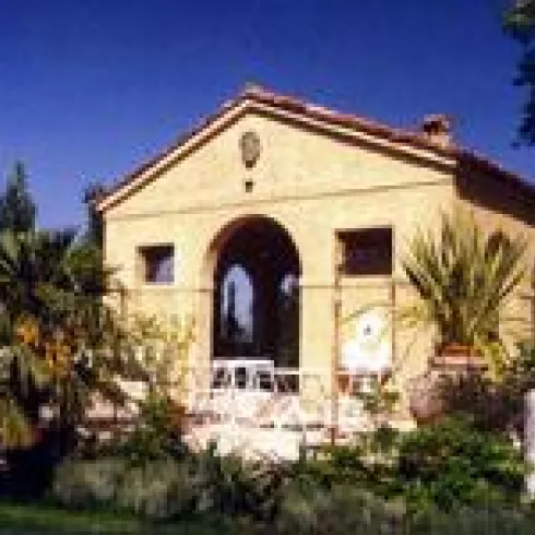 Ein charmantes Haus mit einer sonnigen Veranda und tropischen Pflanzen. Es steht in einer grünen Umgebung mit klarem Himmel.