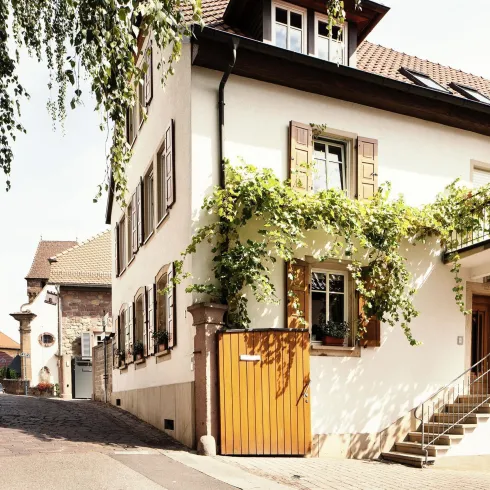 Ein charmantes Haus mit heraushängenden Fensterläden, umgeben von einer malerischen Gasse. Grüne Pflanzen ranken an der Fassade und verleihen dem Ort eine einladende Atmosphäre.