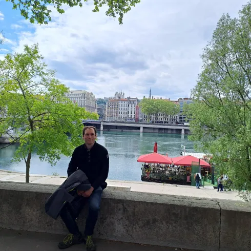 Ein Mann sitzt an einem Flussufer, umgeben von Bäumen. Im Hintergrund sind Gebäude und Stände mit roten Sonnenschirmen zu sehen.