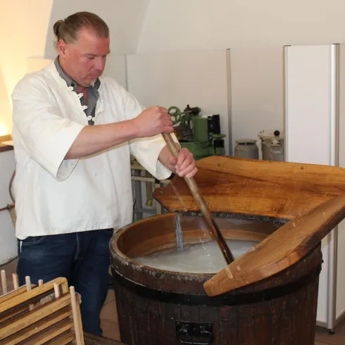 Ein Mann rührt in einem großen Holzfass. Die Szene zeigt traditionelle Handwerkskunst in einer historischen Umgebung.