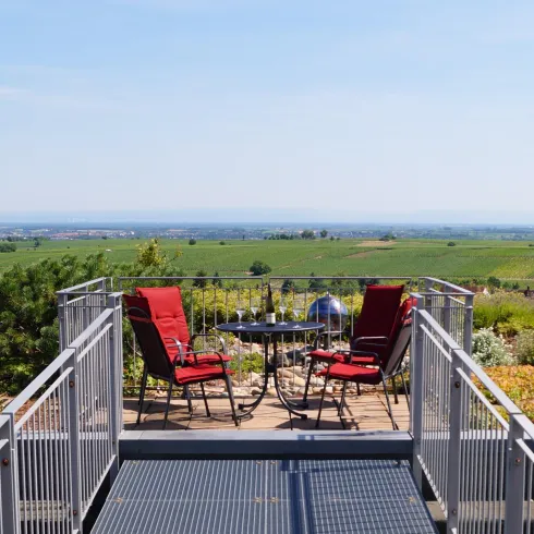 Eine Terrasse mit rot gepolsterten Stühlen und einem kleinen Tisch. Im Hintergrund erstreckt sich eine weite Landschaft mit Weinbergen und klarem Himmel.