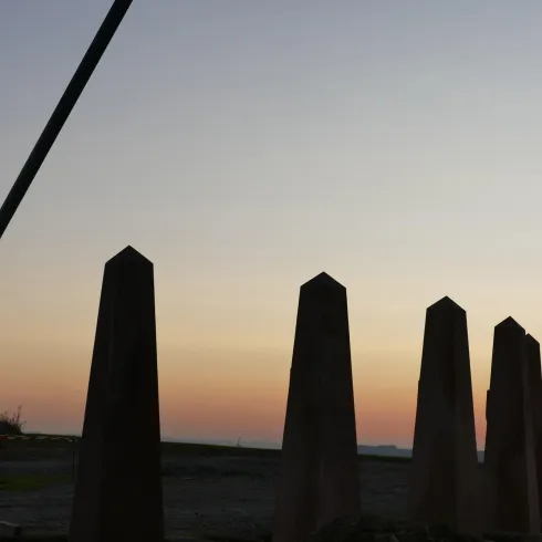 Eine Gruppe von monumentalen Stelen liegt im Vordergrund. Im Hintergrund sieht man den Sonnenuntergang und einen Turm.