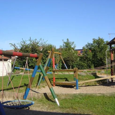Ein Spielplatz mit Schaukeln, einer Rutsche und einer Kletterstruktur. Der Bereich ist von grünen Pflanzen umgeben und hat einen klaren blauen Himmel.